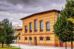 Das Bild zeigt das Gebäude 1 auf dem HSD-Campus, ein zweigeschossiges Backsteingebäude im historischen Industrie-Stil. Charakteristisch sind die großen, hohen Rundbogenfenster sowie das auffällige Ziegelmauerwerk in rot-gelben Farbtönen. Vor dem Gebäude befinden sich eine gepflasterte Fläche mit Bäumen und Hinweisschildern. Der Himmel ist bewölkt und wirkt dramatisch, was dem Bild eine besondere Stimmung verleiht.